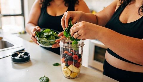 Preparing berry spinach smoothie in sunlit kitchen by two women wearing black activewear