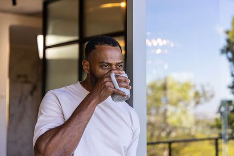 Middle-aged african american man sipping coffee on balcony, gazing through glass door