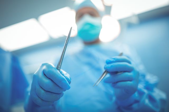 Surgeon holding surgical tools in a sterile operation theater, wearing blue scrubs, gloves, and a mask. Ideal for use in medical articles, healthcare websites, hospital brochures, and educational materials about surgery and medical procedures.