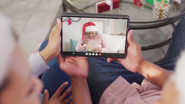 Biracial couple sitting together using tablet for video call with female friend wearing Santa hat. Perfect for themes about holiday communication, virtual celebrations, connecting with family and friends, and festive traditions. This can be used in articles, blogs, promotional materials, and social media posts focusing on modern holiday interactions and maintaining relationships during festivities.