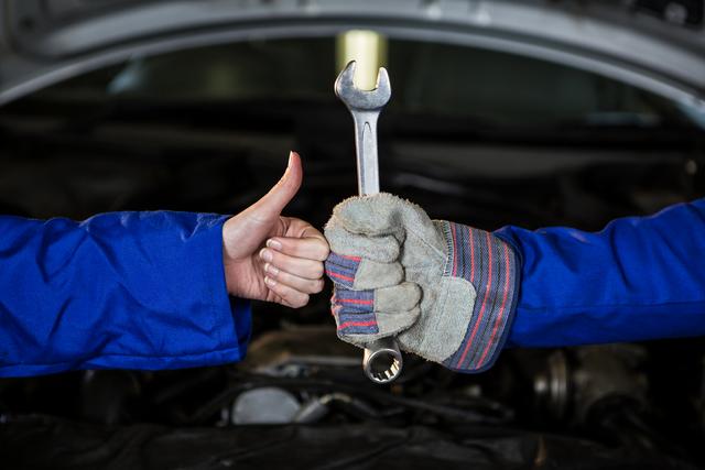 Mechanics in blue uniforms holding a spanner and showing thumbs up, symbolizing successful teamwork and collaboration in an automotive repair garage. Ideal for use in content related to car repair services, mechanical workshops, professional maintenance, and teamwork in the automotive industry.