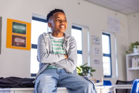 Confident african american boy sitting on desk in bright sunlit classroom