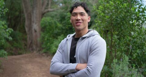 Confident young asian man smiling with arms crossed on forest trail wearing light-gray hoodie