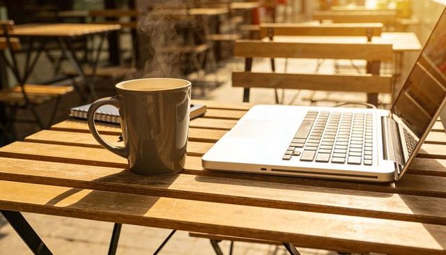 Sunlit outdoor cafe workspace with steaming coffee mug and laptop for remote work