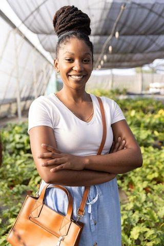 African american woman standing in greenhouse smiling while carrying tan leather handbag