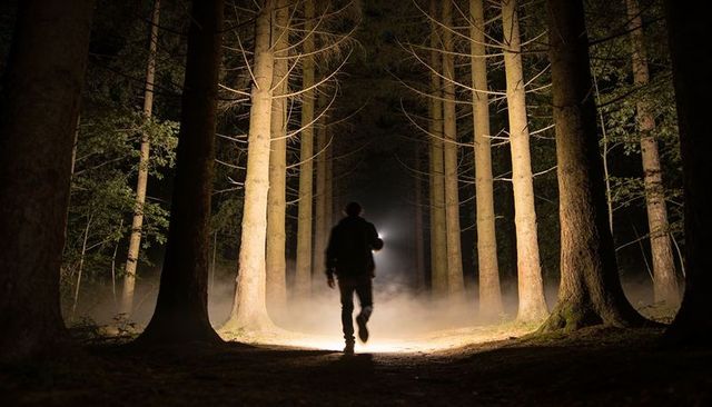 Silhouetted man walking with flashlight on foggy forest trail at night forming mist tunnel
