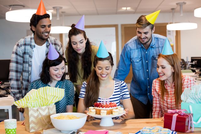Diverse group of colleagues celebrating a birthday in an office environment. They are wearing party hats and smiling while presenting a cake with candles. There are gifts and snacks on the table, creating a festive atmosphere. Ideal for use in articles or advertisements about workplace culture, team building, or office celebrations.