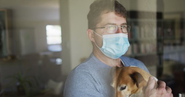 Man standing indoors wearing protective face mask while holding his dog. Looking pensively outside, suggesting a time of reflection and care during the COVID-19 pandemic. Ideal for depicting themes of quarantine, self-care, emotional connection with pets, mental health during pandemic, and public health awareness.