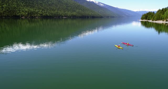 Aerial view of a tranquil mountain lake with two people kayaking. The water is calm, reflecting the surrounding mountains and forests. This image is perfect for use in outdoor travel magazines, adventure blogs, tourism brochures, and environmental campaigns promoting outdoor activities.