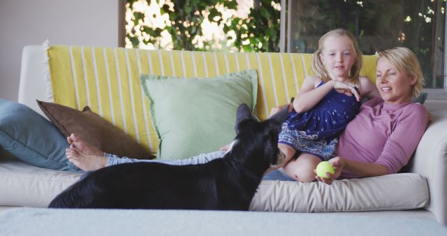 Mother and young daughter are relaxing on the couch at home. Their dog is standing nearby, looking at them. The casual and cozy setting represents quality family time, bonding, and joyful moments. This visual is perfect for advertisements, articles, or social media posts focusing on family life, pet ownership, home living, warmth, affection, and family leisure activities. Ideal for parenting blogs, lifestyle magazines, and promotional materials for home furnishings or pet products.