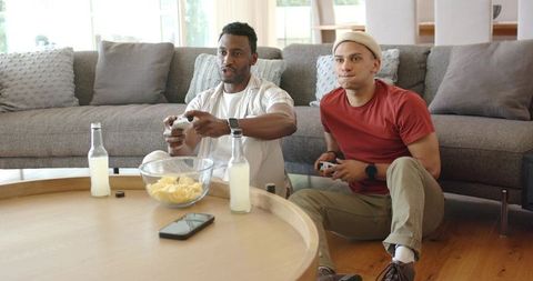 Diverse male friends playing video games on living room floor with snacks and drinks