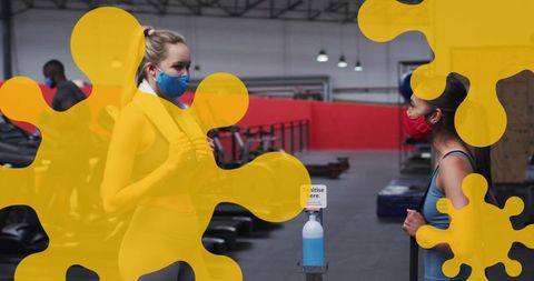 Two women wearing masks chatting in gym beside sanitizer dispenser, demonstrating safety protocols