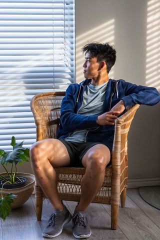 Young asian man sitting in wicker armchair gazing through sunlit blinds