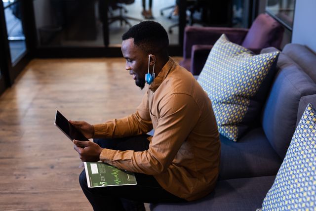 Man sitting on couch in modern office using digital tablet, with face mask hanging from ear. Ideal for illustrating remote work, business continuity during pandemic, health and hygiene practices in workplace, and technology use in professional settings.