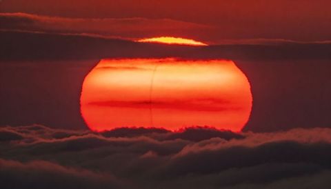 Massive banding sun sinking through layered clouds at fiery amber twilight horizon