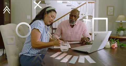 Couple reviewing finances at home dining table with laptop, bills and notepad