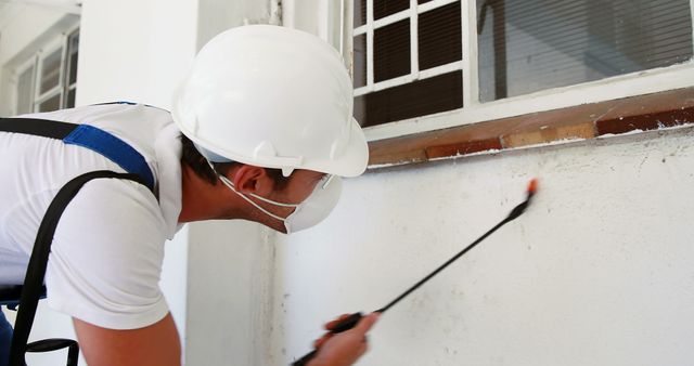 Professional pest control technician wearing protective gear including mask and hard hat, spraying insecticide indoors on a white wall near window. Ideal for content related to pest control services, home maintenance, health and safety precautions, and professional services.