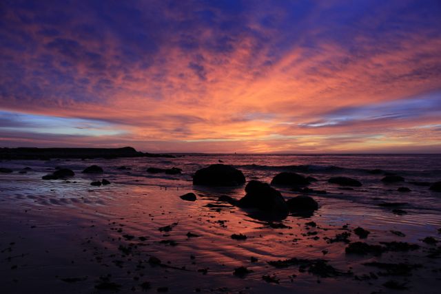 Beautiful sunset over a rocky beach, displaying vibrant colors as the sky reflects on the water and wet sand. Perfect for depicting tranquil scenes, twilight moments, nature's beauty, and serene oceanscapes. Ideal for background images, travel inspiration, environmental themes, and meditation visuals.