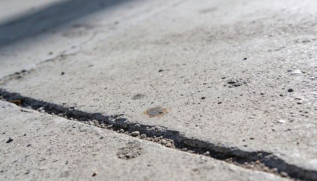 Sunlit Concrete Sidewalk With Diagonal Joint, Gravel-Filled Crack and Rust Stain Closeup Texture