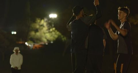 Floodlit night cricket team celebrating bowler wicket with high fives on pitch