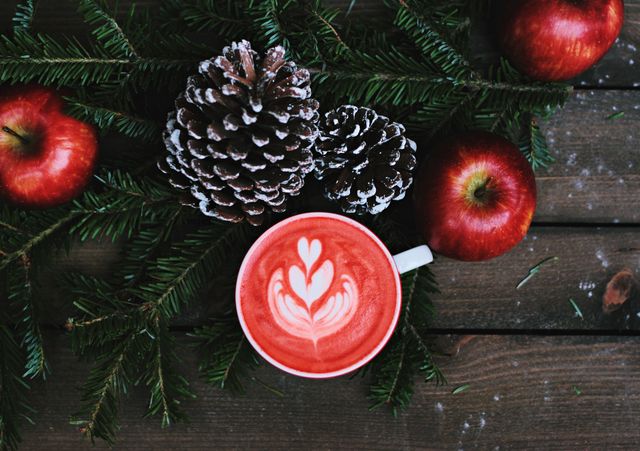 Vibrant festive scene featuring a red latte with beautiful latte art, surrounded by assorted pinecones, fresh red apples, and evergreen boughs on a rustic wooden table. Perfect for use in holiday decorations, coffee shop advertising, winter social media promotions, and seasonal greeting cards.