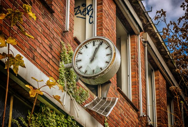 Old clock displaying 11:11 mounted on a brick building with visible graffiti and overgrown vegetation. Overgrown ivy and autumn leaves add a touch of nature reclaiming the space. Ideal for themes showcasing time, urban decay, historical structures, nostalgia, and abandoned places. Suitable for use in articles, presentations, and projects focusing on vintage architecture, urban exploration, and history.
