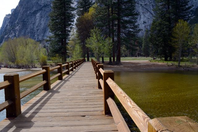 Rustic wooden bridge stretches over calm lake surrounded by dense forest and towering mountains. Ideal for nature blogs, travel promotions, outdoor activity guides.