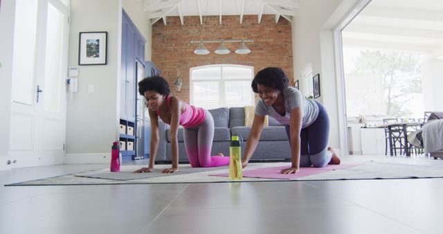 Two young women practicing yoga indoors in a spacious, modern living room. Both are on pink and gray yoga mats, engaging in a stretching pose. They appear focused and relaxed, and there are water bottles beside them indicating they stay hydrated. This image can be used for promotions and articles on indoor fitness, healthy lifestyles, home workout inspirations, and yoga routines.