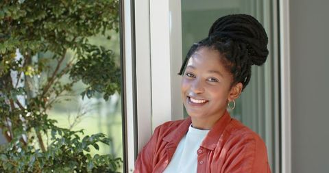 Smiling woman leaning by sliding glass door, folding arms, rust shirt, locs