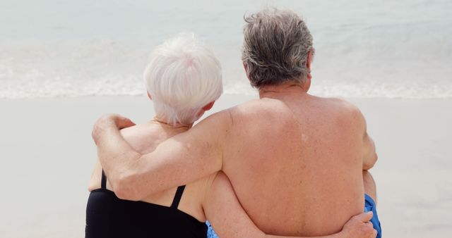 Senior couple sits together on sandy beach, enjoying tranquil ocean view. Ideal for use in content about retirement, senior lifestyle, love and relationships among the elderly, peaceful vacations, and tranquil moments in nature.