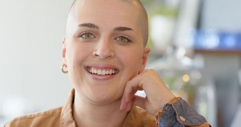 Confident shaved-head woman smiling in cozy cafe with warm bokeh, casual vibe with nose ring