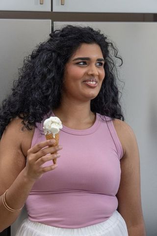 Young woman holding ice cream cone wearing pink tank and gold bracelet in modern kitchen