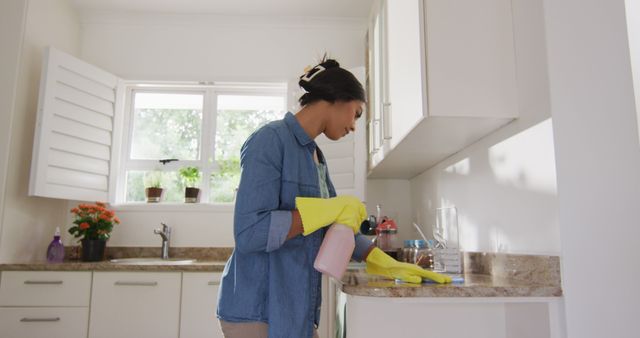 This image shows a woman in yellow gloves cleaning a kitchen counter with a spray bottle. The kitchen is bright and modern, with natural light streaming in through a window with shutters. There are potted plants on the windowsill. This stock photo can be used for articles and advertisements related to home cleaning products, domestic chores, household hygiene, and promoting a clean living environment.