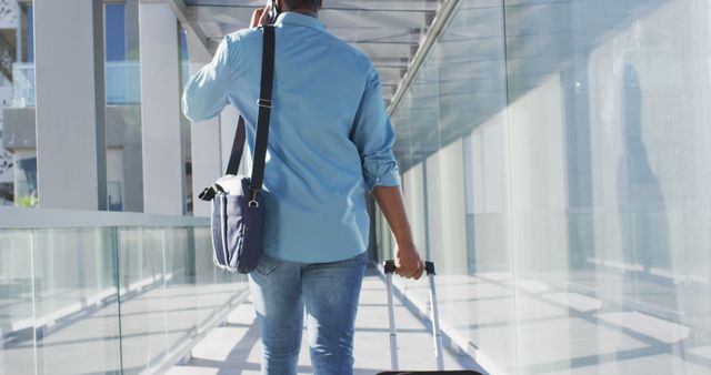 Business traveler wearing casual wear including a blue shirt and jeans, walking through a modern airport corridor with luggage. Ideal for concepts related to business travel, tourism, work trips, transportation, and urban lifestyle. Can be used in promotional content for airlines, travel agencies, and business travel services.