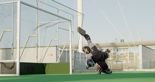 Side view of a Caucasian male field hockey goalkeeper, diving to stop the ball with his hockey stick during the game on the hockey pitch, with his teammates and opponents in the background