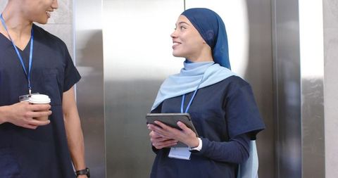 Hospital staff chatting in elevator, nurse in hijab holding tablet and wearing badge