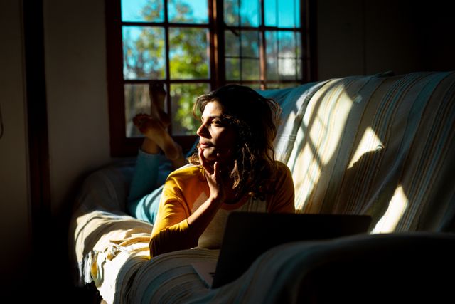 A relaxed biracial woman is lying on a sofa using a laptop in a sunny cottage living room. The scene is filled with natural light, creating a cozy and serene atmosphere. This image can be used for promoting healthy living, remote work, home office setups, and lifestyle blogs. It is also suitable for advertisements related to technology, leisure, and rural home living.