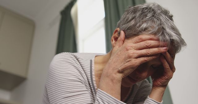 Elderly woman with short gray hair holding her head in distress, likely experiencing headache, stress, or mental health issues, in a domestic setting with gray curtains in the background. Useful for depicting mental health, senior citizen issues, and emotional struggles in older adults.