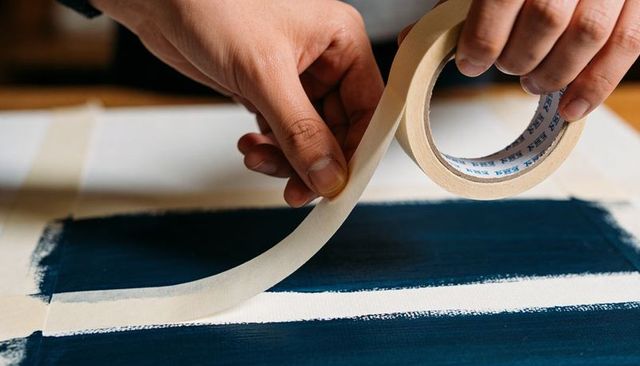 Hands peeling masking tape revealing crisp white stripe on navy blue painted panel closeup