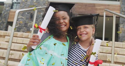 Joyful graduates wearing mortarboards holding diplomas with confetti falling on steps