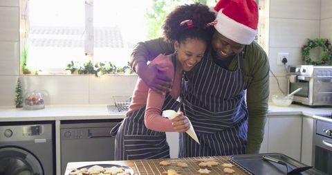 African american couple decorating christmas cookies in cozy kitchen wearing santa hat