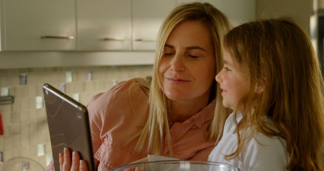 A mother and her young daughter are using a tablet while cooking together in a modern kitchen. This image can be used to depict family bonding, modern kitchen activities, or technology use in everyday life. Ideal for websites, blogs, advertisements, and social media relating to family life, home cooking, or digital technology.