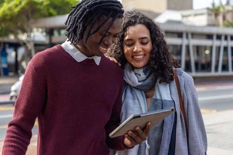 Smiling diverse couple walking city street browsing tablet, urban tech lifestyle