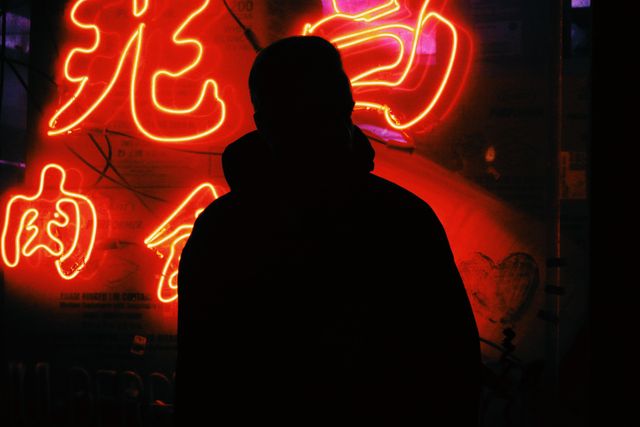 Silhouette of an individual standing in front of brightly lit neon signs at night. Red and orange hues create a vibrant urban atmosphere. Ideal for themes involving nightlife, city life, and anonymous moods. Perfect for use in blogs, websites, promotional materials, or artistic projects focusing on urban exploration or nightlife culture.