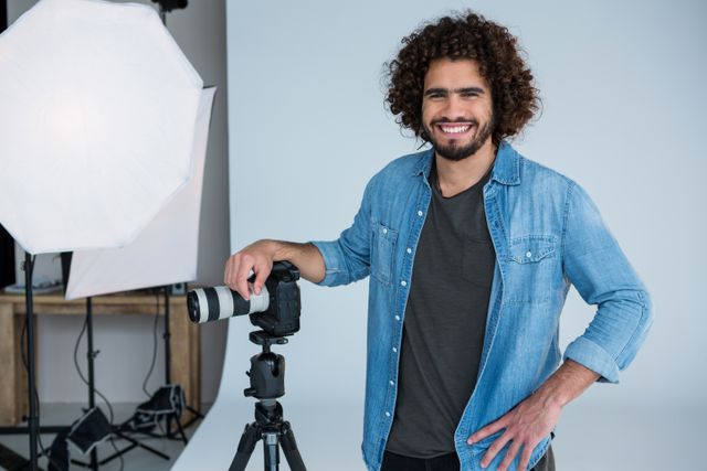 This image shows a smiling male photographer standing in a studio with his camera on a tripod. He is wearing a denim shirt and has curly hair. The studio is equipped with lighting equipment, indicating a professional photography setting. This image can be used for articles or advertisements related to photography, creative professions, studio setups, or professional portraits.