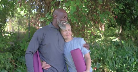 Mature diverse couple embracing after outdoor yoga, holding purple yoga mats, smiling