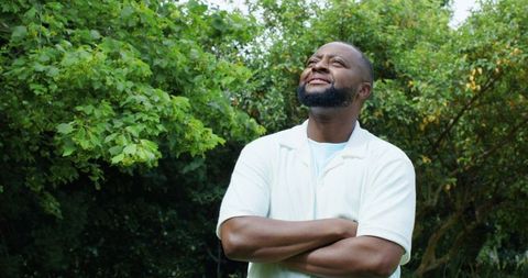 Man standing arms crossed looking up smiling in garden with lush green foliage, serene confidence