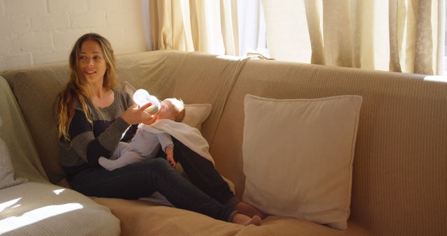 A Caucasian young woman is sitting on a couch, feeding a baby with a bottle, with copy space. Her relaxed posture and gentle interaction with the child convey a sense of maternal care and bonding.