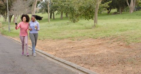 Diverse women walking together in park wearing activewear, holding phone and water bottle