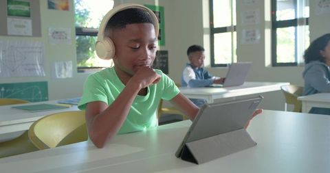 Elementary student using tablet with headphones in bright classroom streaming lesson